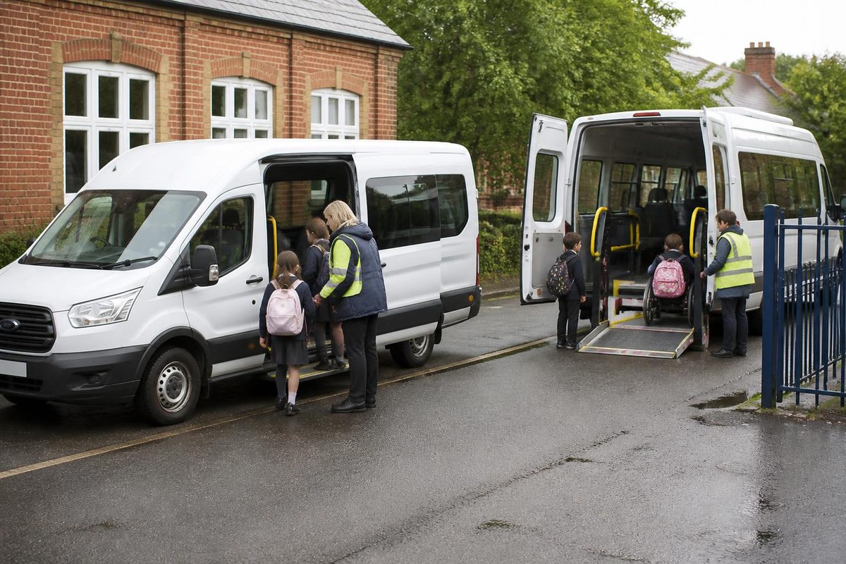 Silver people carrier with its side door open outside a primary school gate as a passenger assistant waits on the pavement